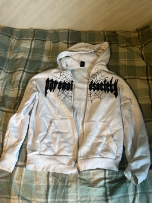 Vit hoodie med tryck och dragkedja - Vit hoodie med svart tryck i gotisk stil och spindelnätsdetaljer på bröstet. Modellen har huva, dragkedja framtill och två fickor. Perfekt för dig som gillar streetwear och vill sticka ut med en unik design.