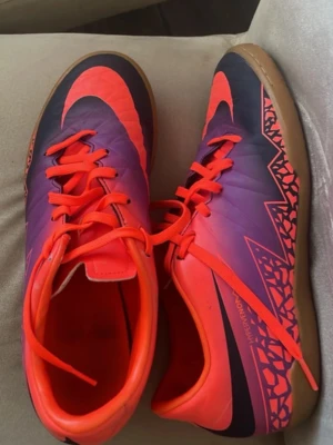Nike fotbollsskor inomhus lila/orange - Säljer ett par Nike inomhusfotbollsskor i lila och orange med grafiskt mönster på sidan och gummisula. Skorna har snörning och en låg profil som passar perfekt för snabba rörelser på planen. Snygg design med stora Nike-loggor och bekväm passform.