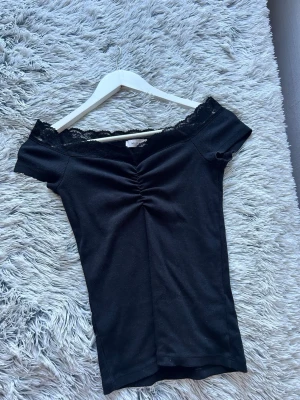 Svart offshoulder topp med spets - Snygg svart offshoulder topp med spetskant upptill och rynkdetalj framtill. Köpt från Nelly och är i storlek M, men skulle säga att det passar en S 💕(säljer för den aldrig kommer till användning, + använt endast 1 gång)  