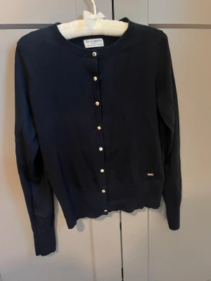 Mörkblå kofta från Holly & White - Klassisk mörkblå kofta från Holly & White med rund halsringning och pärlemorskimrande knappar framtill. Långärmad modell i mjukt material som passar perfekt till lager på lager. Enkel och stilren design med liten logodetalj vid nederkanten.