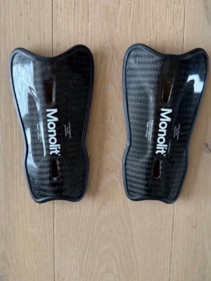 Svarta benskydd från Monolit - Säljer ett par svarta benskydd från Monolit i kolfiber-look. De har en ergonomisk form och två ventilationshål för extra komfort. Perfekta för dig som vill ha lätta och tåliga skydd till fotboll eller annan sport.