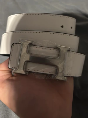 Vitt bälte med stort H-spänne - Snyggt vitt bälte med ett stort, blankt H-format spänne i silverfärg. Bältet har stilrena svarta sömmar längs kanterna och är gjort i slätt material som ger en clean look. Perfekt accessoar för att lyfta vilken outfit som helst. Endast bälte+ hole