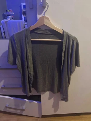 Grå kortärmad öppen kofta - En enkel och tunn grå kofta med korta ärmar och öppen framtill. Perfekt att slänga över ett linne eller t-shirt för en avslappnad look. Materialet är mjukt och stretchigt, vilket gör den bekväm att bära. I storlek M men passar även som en S