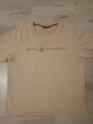 t-shirt från Sail Racing - Snygg beige t-shirt från Sail Racing med tryckt logga på bröstet. Klassisk rund halsringning och korta ärmar. Tillverkad i mjuk bomull som känns skön mot huden. Perfekt för en avslappnad och stilren look.