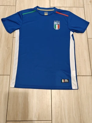 Blå Italien fotbollströja - Snygg blå fotbollströja med Italiens landslagsemblem på bröstet och texten 'ITALIA' på ryggen. Tröjan har vita sidopaneler och detaljer i grönt och rött på axlarna. Tillverkad i ett lätt och ventilerande material, perfekt för träning eller match.