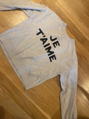 Grå sweatshirt med text JE T'AIME - Ljusgrå sweatshirt med svart tryck 'JE T'AIME' på bröstet. Tröjan har rund halsringning och långa ärmar. Perfekt för en avslappnad och trendig look. Materialet känns mjukt och bekvämt.
