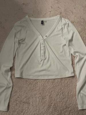 Vit topp med knappar - Vit croppad topp med ribbad struktur och v-ringning i storlek M. Toppen har långa ärmar och knappar framtill för en snygg detalj. Perfekt att matcha med höga jeans eller kjol för en trendig look. Aldrig använd🩷