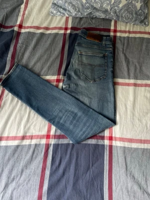 Blå slim fit jeans  - Säljer ett par blå slim fit jeans från Tiger of Sweden i modellen EVOLVE med klassisk tvätt och snygga slitningar. Jeansen har ett litet hål på vänstra knät men det påverkar inte helhetsintrycket. Perfekta till en avslappnad look och passar till det mesta i garderoben. Hör gärna av er om ni har några frågor eller funderingar! 