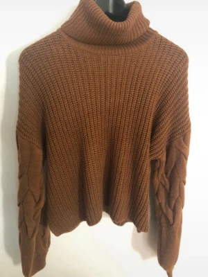 Brun polotröja med flätade ärmar - Säljer en chunky brun stickad polotröja med bred ribbstickning och snygga flätade detaljer längs ärmarna. Tröjan har en hög polokrage och avslappnad passform, perfekt för kalla dagar. Materialet känns mjukt och värmande. Kort i modellen