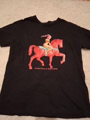 Svart t-shirt med tryck Fröken snusk  - Cool svart t-shirt med färgstarkt tryck av en rosa häst och en tecknad figur med texten 'Rasmus Gozzi & Fröken Snusk' under motivet. T-shirten har rund hals och korta ärmar. Materialet samlar på sig hår ganska lätt men inget mer än så, annars väldigt bra skick