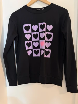 Svart tröja med lila hjärtan - Cool svart sweatshirt med lång ärm och tryck av lila rutor med svarta hjärtan på framsidan. Tröjan har rund halsringning och ribbade muddar vid ärmslut och nederkant. Har använt 2-3 gånger.