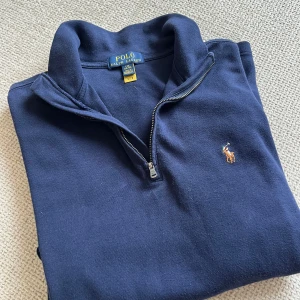 Mörkblå quarter zip från Polo Ralph Lauren - kille  - Klassisk mörkblå quarter zip-tröja från Polo Ralph Lauren med broderad logga på bröstet. Tröjan har hög krage, dragkedja framtill och långa ärmar. Perfekt för en avslappnad men ändå stilren look. Storlek junior. 