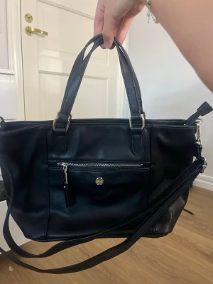 Svart handväska med axelrem - Aldrig använd svart handväska i konstläder med både handtag och avtagbar axelrem. Väskan har silverfärgade detaljer, två ytterfack med dragkedja och en enkel, klassisk form. Ursprungpriset 500-900 köpt för länge sedan kommer ej ihåg.
