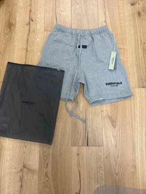 Grå Essentials shorts med snörning - Snygga grå shorts från Essentials med elastisk midja och snörning framtill. Klassisk passform och diskret logga på vänster ben. Perfekta för chill dagar eller träning. Tillverkade i mjuk bomull för maximal komfort.