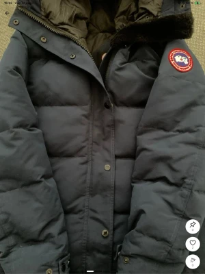 Mörkblå dunjacka från Canada Goose - Säljer en mörkblå dunjacka från Canada Goose med klassisk patch på ärmen. Jackan har huva med mjuk pälsdetalj, dragkedja och knappar framtill samt quiltad design för extra värme. Perfekt för kalla vinterdagar.
