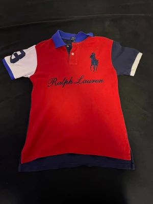 Röd och marinblå pikétröja Ralph Lauren - Snygg pikétröja från Ralph Lauren i rött och marinblått med blå krage och kontrasterande ärmar i vitt och blått. Stor logga och text på bröstet samt nummerdetalj på ena ärmen. Klassisk polokrage och knappar framtill.