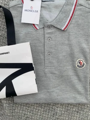 Grå pikétröja från Moncler - Snygg grå pikétröja från Moncler med klassisk krage och tre knappar framtill. Kragen har röda och vita ränder som ger en sportig touch. Moncler-logga är broderad på bröstet. Perfekt till både jeans och shorts.