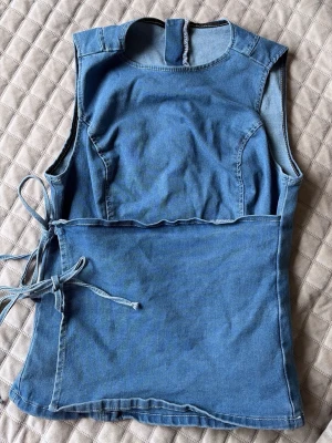 Ärmlös denim korsettopp med knyt - Säljer en snygg ärmlös topp i blå denim med korsettliknande passform och knytband i sidan. Toppen har rund halsringning och dragkedja i ryggen för enkel påklädning. Perfekt för dig som vill ha en trendig och unik look. Från SHEIN. 