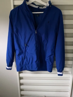 Ralph Lauren Windbreaker - Snygg blå vindjacka med dragkedja framtill och två fickor med dragkedja. Jackan har huva och vita detaljer på muddarna. Perfekt för blåsiga dagar och enkel att matcha med olika outfits.