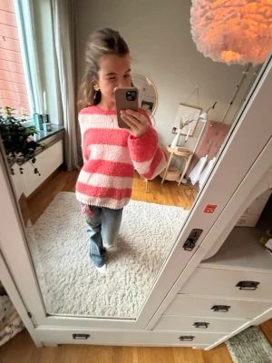 Rosa och vit randig stickad tröja - Mysig stickad tröja med breda rosa och vita ränder. Tröjan har en avslappnad passform, rund halsringning och långa ärmar. Perfekt för att hålla sig varm och snygg under kyliga dagar.