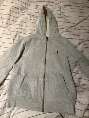 Grå hoodie från Polo Ralph Lauren - Klassisk grå hoodie från Polo Ralph Lauren med dragkedja framtill och två fickor. Broderad logga på bröstet och mjukt, fleecefodrat material. Perfekt för en avslappnad och stilren look. Huva med snörning för extra komfort.