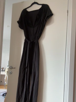 Svart jumpsuit med spetsdetaljer - Snygg svart jumpsuit med vida ben och korta ärmar. Djup v-ringning med dekorativa spetsdetaljer runt halsen och knytband i midjan. Tillverkad i ett mjukt, tunt tyg som ger en avslappnad look.