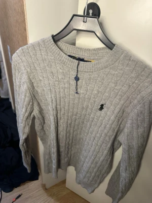 Ljusgrå kabelstickad tröja Polo - Ljusgrå kabelstickad tröja från Polo Ralph Lauren med rund halsringning och den klassiska broderade loggan på bröstet. Tröjan har långa ärmar och ribbade muddar vid ärmslut och nederkant. Perfekt för lager på lager och stilren vardagslook.