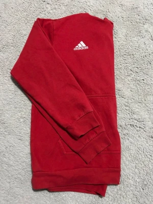 Röd hoodie från Adidas - Snygg röd hoodie från Adidas med vit logga på bröstet. Klassisk känguruficka framtill och huva med vita dragsnören. Tillverkad i mjukt sweatshirtmaterial, perfekt för en avslappnad stil.