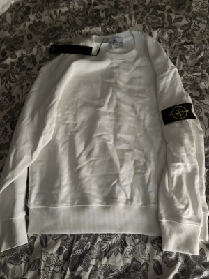 Vit sweatshirt från Stone Island - Säljer en stilren vit sweatshirt från Stone Island med den klassiska svarta logotypen på vänster ärm. Tröjan har rund halsringning, ribbade muddar och är tillverkad i mjukt bomullsmaterial. Perfekt för en clean och modern look.