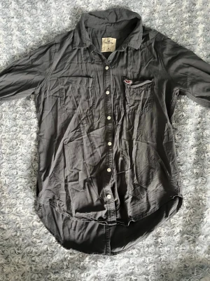 Mörkgrå skjorta från Hollister - Snygg mörkgrå skjorta från Hollister med klassisk krage, två bröstfickor och vita knappar framtill. Skjortan har lång ärm och en liten Hollister-logga på ena fickan. Perfekt för en avslappnad och stilren look. Den har legat vikt därav att den är skrynklig men går att stryka:)