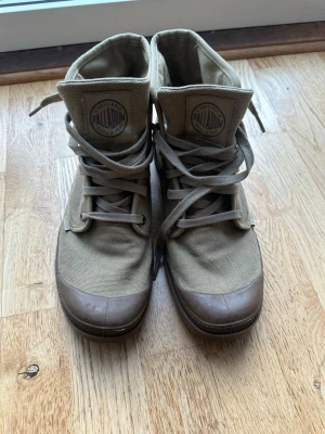 Beige canvas kängor från Palladium - Säljer ett par beige höga kängor från Palladium i slitstark canvas med gummiförstärkt tå och robust sula. Klassisk logga på plösen och snörning hela vägen upp. Perfekta för dig som gillar streetwear och vill ha en stilren men tuff look.