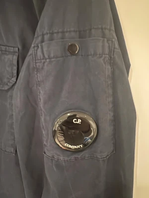Mörkblå overshirt från C.P. Company - Snygg mörkblå overshirt från C.P. Company med ikonisk linsdetalj på ärmen och knappstängning. Overshirten har en klassisk krage och ficka på ärmen. Perfekt för lager-på-lager och streetwear-stil.