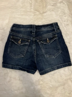 Mörkblå denimshorts med fickor - Snygga mörkblå denimshorts med klassisk femficksdesign och kontrastsömmar. Shortsen har bälteshällor, knapp- och dragkedjestängning samt dekorativa ficklock med knapp bak. Perfekta för varma dagar och enkel att matcha med olika toppar. Ljusterbar i midjan.