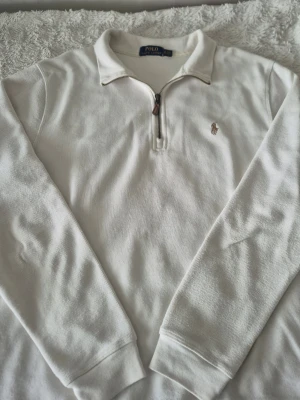 Polo Ralph Lauren quarter zip - Klassisk vit långärmad polotopp från Polo Ralph Lauren med half zip och broderad logga på bröstet. Tillverkad i mjuk bomull med ribbade muddar och hög krage. Perfekt för en clean och stilren look.