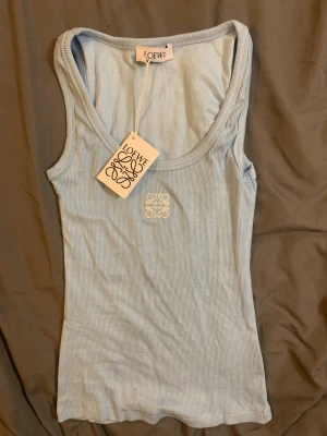 Ljusblå ribbad tanktop från Loewe - Supersnygg ljusblå ribbad tanktop från Loewe med broderad logga framtill. Stretchigt material och breda axelband ger en clean och trendig look. Perfekt att styla med jeans eller kjol för en fräsch vibe.