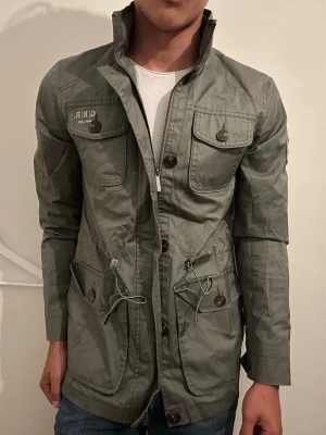  Field jacket med patch och snörning - Stilren grön vindjacka med hög krage, flera fickor med knappar och snörning i midjan. Jackan har patch med brittisk flagga på ärmen och tryck på bröstfickan. Perfekt för vår och höst. Grön grå väldigt unik