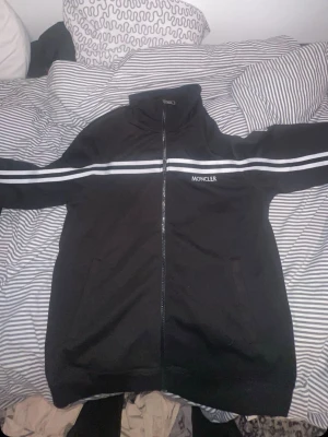 Svart zip hoodie från Moncler - Säljer en svart hoodie från Moncler med dragkedja framtill och vita dubbla ränder över bröstet och armarna. Moncler-loggan är broderad på bröstet. Hoodien har hög krage och två fickor framtill. Materialet känns mjukt och bekvämt.