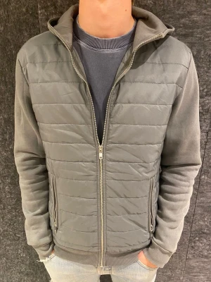 Grå hoodie med dragkedja och huva - Snygg grå hoodie med quiltad framsida, dragkedja och två fickor med dragkedja. Mjuk bomullsmix i ärmar och rygg, samt en skön huva. Perfekt för en avslappnad och stilren look.