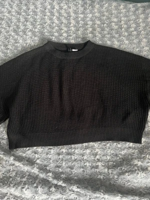 Svart cropped stickad tröja - Säljer en svart cropped stickad tröja med korta ärmar och ribbad struktur. Tröjan har en högre rund halsringning och bred mudd nedtill. Perfekt att styla över en skjorta eller med höga jeans.