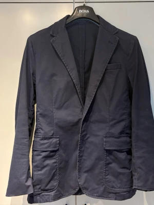 Mörkblå kavaj med fickor - Stilren mörkblå kavaj med två stora fickor framtill och en bröstficka. Klassisk passform med enkelknäppning och slits bak. Kavajen är tvättbar i maskin och får även strykas. Nypris ligger på ca 3300. Den är använd sparsamt och nytvättad😊