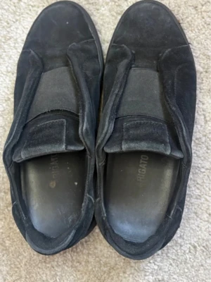 Svarta loafers från Axel Arigato - Säljer ett par svarta loafers från Axel Arigato i mocka med klassisk slip-on design. Skorna har en stilren look med breda elastiska band över vristen och robust gummisula med mönster för bra grepp. Perfekta för dig som gillar minimalistisk och modern stil. De är ganska sparsamt använda och har inga slitningar eller skador. 