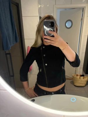 Svart jeansjacka med dragkedjor - Snygg svart jeansjacka med asymmetrisk dragkedja och silverfärgade knappar. Jackan har två dragkedjefickor framtill och en kort, figurnära passform. Perfekt för en trendig och cool look.