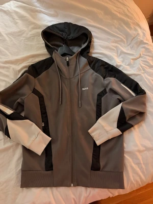 Grå hoodie med dragkedja från BOSS - Snygg grå hoodie från BOSS med dragkedja framtill, svarta och vita detaljer på ärmar och axlar samt justerbar huva med snören. Tröjan har ribbade muddar och två fickor med dragkedja. Perfekt för en sportig och avslappnad stil.