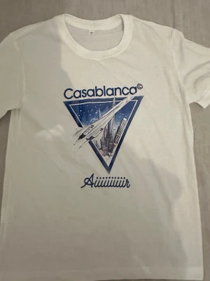 Vit Casablanca t-shirt med tryck - Vit t-shirt med blått och vitt tryck från Casablanca. Framsidan har ett grafiskt motiv med ett flygplan, stadssilhuett och texten 'Casablanca' samt 'Aiiiiiiiir'. Klassisk rund hals och korta ärmar. Perfekt för en cool och avslappnad stil.