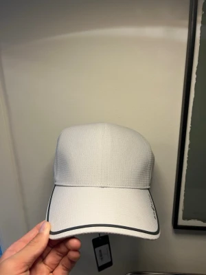 Vit keps från Sail Racing - Helt ny snygg vit keps från Sail Racing med diskret logga på sidan av skärmen. Kepsen har en sportig look med grå kant runt skärmen och är tillverkad i ett lätt, ventilerande material med struktur. Perfekt för dig som gillar stilren och funktionell accessoar. 