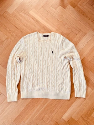 Polo Ralph Lauren kabelstickad Tröja Cream M - Kabelstickad tröja i krämvitt från Polo Ralph Lauren med rund halsringning och broderad marinblå logga på bröstet. Tröjan har ribbade muddar vid ärmslut och nederkant. Perfekt för en klassisk och stilren look.