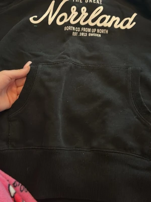 Svart hoodie The Great Norrland - Säljer en svart hoodie med vit text 'The Great Norrland' och detaljer från SQRTN Co. på bröstet. Klassisk känguruficka framtill och ribbade muddar. Perfekt för dig som gillar streetwear och vill ha något bekvämt och stilrent.