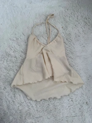 Ljusgul omlott linnetopp med knyt - Säljer en ljusbeige linnetopp med omlottdesign och knyt i nacken. Toppen har en lätt volangkant nedtill och är ärmlös med v-ringad hals. Perfekt för sommardagar och ger en luftig känsla.