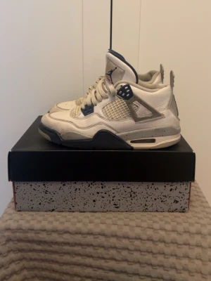 Vita och marinblå Air Jordan 4 - Säljer ett par Air Jordan 4 sneakers i vitt och marinblått med meshpaneler och detaljer i plast. Skorna har den klassiska höga siluetten, synlig Air-enhet i sulan och Jumpman-logga på plösen. Perfekta för dig som gillar retrostil och streetwear.