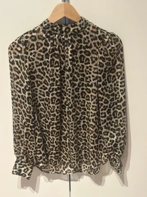 Leopardmönstrad blus med lång ärm - Snygg blus med leopardmönster i beige, brunt och svart. Blusen har lång ärm med knappar vid ärmslut och en hög krage. Tillverkad i ett tunt, lätt material som ger ett luftigt intryck. En av hakarna för att fästa knappen har gått sönder, se sista bilden. 